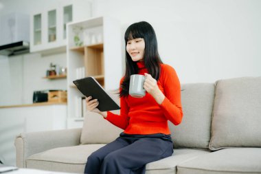 Genç Asyalı öğrenci evdeki kanepede oturup dijital tablet kullanıyor ve kahve içiyor. 