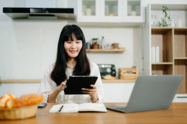 Genç iş kadını mutfakta oturup dizüstü bilgisayar ve dijital tablet üzerinde çalışıyor. 