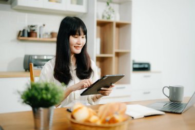 Genç iş kadını mutfakta oturup dizüstü bilgisayar ve dijital tablet üzerinde çalışıyor. 