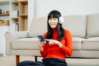 Genç Asyalı kulaklıklı öğrenci oturma odasında yerde oturuyor ve akıllı telefon kullanıyor.  