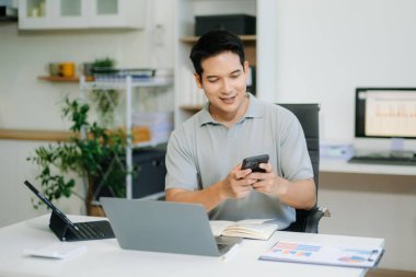 Kendine güvenen iş uzmanı, gülümseyen, dizüstü bilgisayarda yazan ve dijital tableti yaratıcı ofiste tutan çekici genç adam.
