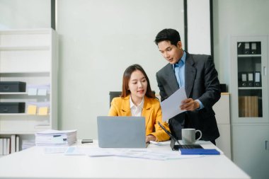 İki Asyalı iş adamı akıllı telefonla konuşuyor ve ofiste dizüstü bilgisayar kullanıyorlar.