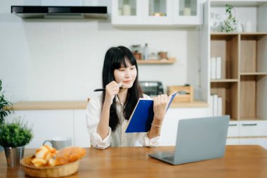 Genç iş kadını mutfakta oturuyor ve dizüstü bilgisayarda çalışıyor ve not defterine not alıyor. 