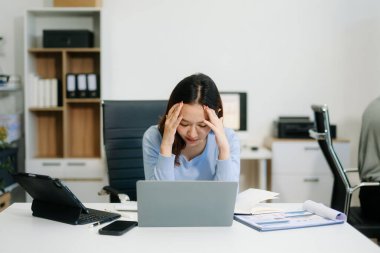Asyalı kadın migren ağrısı çekiyor. Ofiste dizüstü bilgisayar ve tablet üzerinde çalışırken çok çalışan iş kadını finansör.