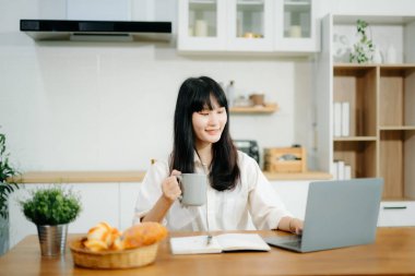Genç iş kadını mutfakta oturuyor ve kahve içerken dizüstü bilgisayarda çalışıyor.