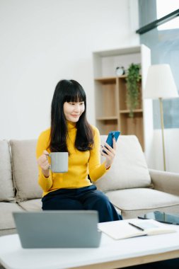 Genç Asyalı öğrenci evde kanepede oturuyor ve kahve içerken bilgisayarla akıllı telefon kullanıyor. 