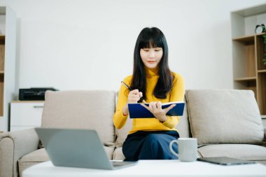 Genç Asyalı öğrenci evdeki kanepede oturuyor, not defterine not alıyor ve dizüstü bilgisayar kullanıyor. 