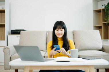 Genç Asyalı öğrenci evdeki kanepede oturuyor ve akıllı telefon kullanıyor. 