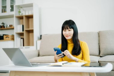 Genç Asyalı öğrenci evdeki kanepede oturuyor ve bilgisayarlı akıllı telefon kullanıyor. 