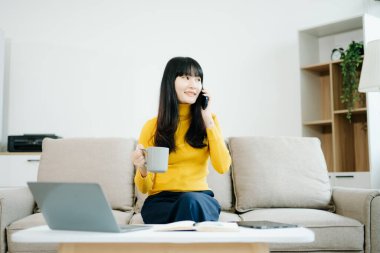 Genç Asyalı öğrenci evdeki kanepede oturuyor, kahve içiyor ve akıllı telefondan konuşuyor. 