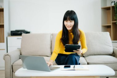 Genç Asyalı öğrenci evdeki kanepede oturuyor ve dizüstü bilgisayar ve dijital tablet kullanıyor. 