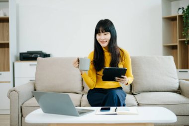 Genç Asyalı öğrenci evdeki kanepede oturup dijital tablet kullanıyor ve kahve içiyor. 