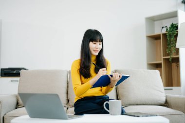 Genç Asyalı öğrenci evdeki kanepede oturuyor, not defterine not alıyor ve dizüstü bilgisayar kullanıyor. 