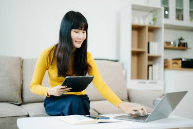 Genç Asyalı öğrenci evdeki kanepede oturuyor ve dijital tabletle dizüstü bilgisayar kullanıyor.  