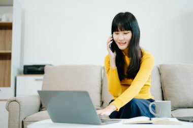 Genç Asyalı öğrenci evinde kanepede oturuyor ve dizüstü bilgisayar kullanıyor, akıllı telefondan konuşuyor. 