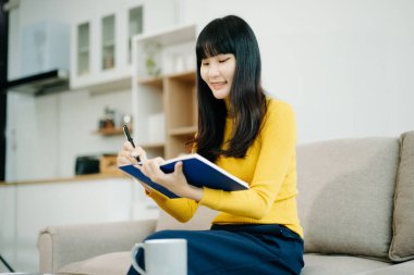 Genç Asyalı öğrenci evdeki kanepede oturuyor ve not defterine not alıyor.  