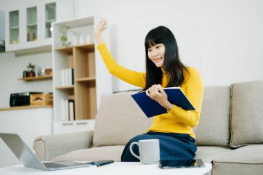 Genç Asyalı öğrenci evdeki kanepede oturuyor ve dizüstü bilgisayar kullanıyor, not defteri tutuyor ve el kol hareketi yapıyor. 