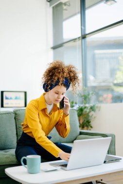 Merkez ofisteki yeşil bir koltukta otururken bilgisayar ve akıllı telefon kullanan bir kadının portresi.