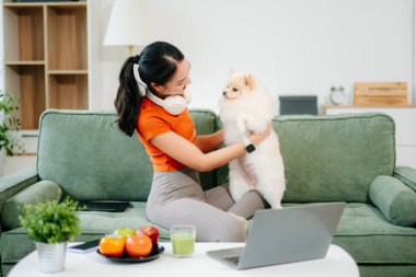 Beyaz kanepede oturan genç bir kadın evdeki parlak, modern oturma odasında küçük beyaz bir köpek yavrusu tutuyor.