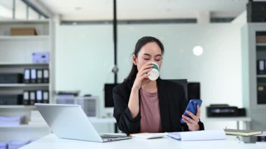 Genç iş kadını masada oturmuş akıllı telefon kullanarak kahve içiyor.