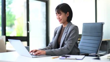 Gri ceketli genç Asyalı iş kadını modern ofiste çalışıyor.