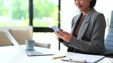 Gri ceketli genç Asyalı iş kadını modern ofiste akıllı telefon kullanıyor.