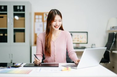 Modern ofiste dijital tablet kalem ve dizüstü bilgisayar kullanan genç bir kadın grafik tasarımcısı. Yaratıcı çalışma alanı, renk paletleri ve UI UX tasarımı devam ediyor