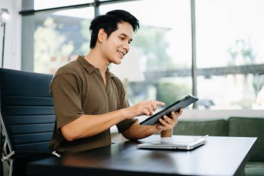 Genç Asyalı adam modern ofis masasında dizüstü bilgisayarda kahve ve tabletle çalışıyor. Teknoloji, serbest çalışma, iş ve uzak çalışma temaları için ideal.