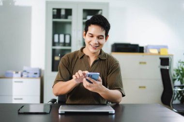 Modern ofis masasında akıllı telefon kullanan mutlu bir Asyalı. Dijital yaşam tarzı, uzak çalışma, iletişim, serbest çalışma kavramları için ideal