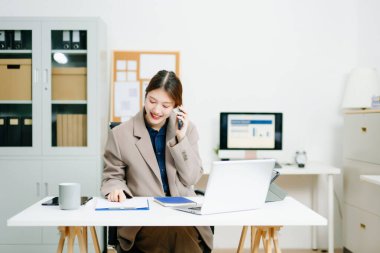 Modern bir ofiste dizüstü bilgisayarla çalışırken gülümseyen bir iş kadını telefonda konuşuyor. İletişim, pazarlama, finans ve dijital çalışma kültürü temaları için mükemmel