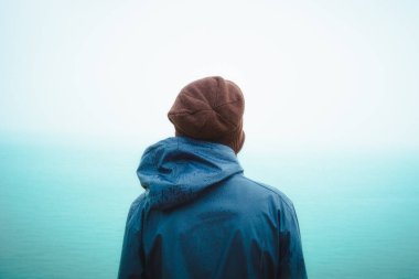 Yağmurluk ve yün şapka giyen bir kişi, yağmurun altında duruyor, arkadan görünüyor, kasvetli ve düşünceli bir atmosferde sisli deniz ufkuna bakıyor..