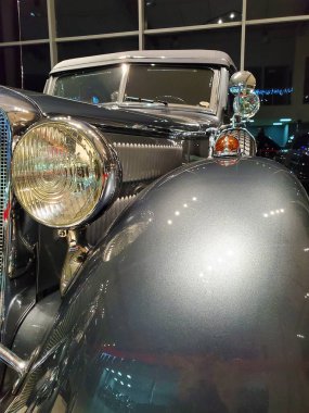 Close-up front view of a vintage classic car with polished chrome details, round headlights, and glossy metallic paint, displayed indoors under artificial lighting. The image highlights the