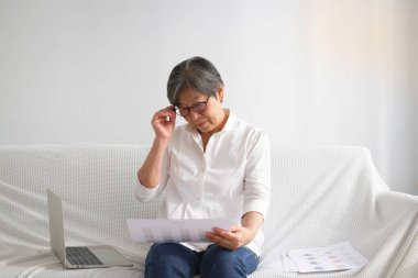 Yaşlı akıllı bayan elinde iş haritası, oturma odasında e-öğrenim için dizüstü bilgisayar kullanıyor, orta yaşlı yaşam tarzı iyi.