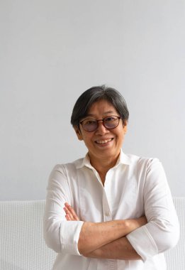 Beyaz gömlek giyen, kolları çapraz duran, gülümseyen, mutlu hisseden, poz veren yaşlı ve zeki bir kadın.