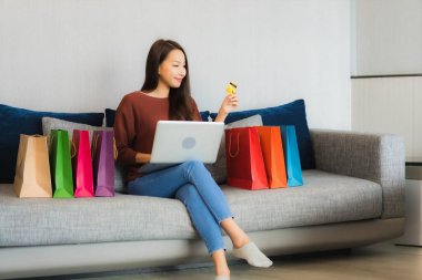 Asyalı güzel bir kadının portresi oturma odasındaki kanepede online alışveriş yapmak için bilgisayar ve kredi kartı kullanıyor.