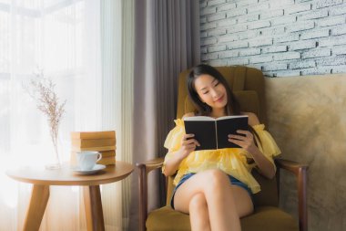 Genç Asyalı kadının portresi kanepeye oturur ve oturma odasının içinde kahve fincanı ile kitap okur.