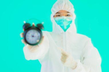 Güzel bir portre genç Asyalı doktor. Özel süit ve maske takıyor. Mavi arka planda saati ya da alarmı gösteriyor.