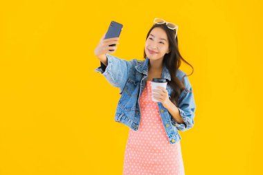 Kahve fincanı ve sarı izole edilmiş arka planda akıllı cep telefonlu güzel Asyalı kadın portresi.