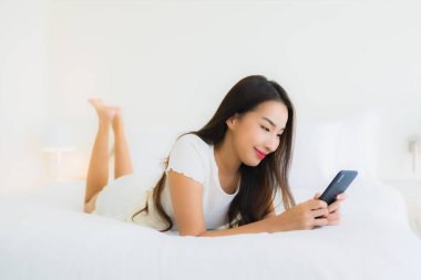 Güzel bir Asyalı kadın portresi. Yatak odasında beyaz yastık battaniyesi ve yatağında akıllı cep telefonu kullanıyor.