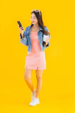 Kahve fincanı ve sarı izole edilmiş arka planda akıllı cep telefonlu güzel Asyalı kadın portresi.