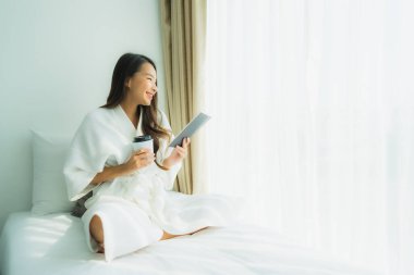 Genç Asyalı kadın, elinde kahve fincanıyla yatak odasında kitap okuyor.