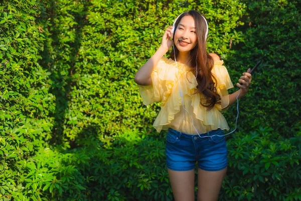 若いアジアの女性は 屋外庭園の自然景観の周りの音楽を聴くためのヘッドフォン付きスマートフォンを使用して — ストック写真