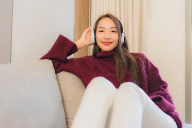 Güzel bir Asyalı kadının portresi oturma odasındaki kanepede müzik dinlemek için kulaklık kullanıyor.