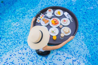 Asyalı güzel bir kadının portresi yüzme havuzunda yüzen kahvaltı tepsisiyle mutlu bir gülümseme