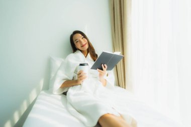 Genç Asyalı kadın, elinde kahve fincanıyla yatak odasında kitap okuyor.