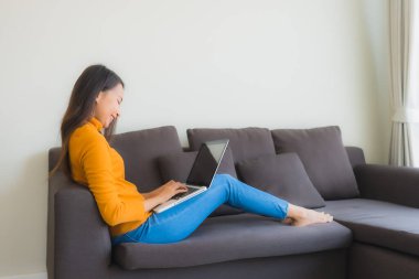 Genç Asyalı bir kadının oturma odasında çalışmak için dizüstü bilgisayar defteri kullanmasının portresi.