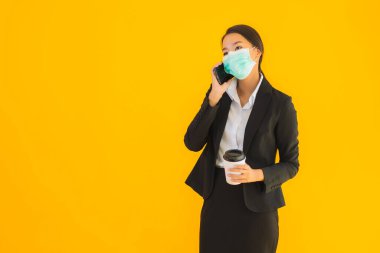 Portre güzel genç Asyalı kadın maske takıyor. Cep telefonu ve kahve ile covid19 ve koronavirüs konseptini sarı izole arka planda kullanın.
