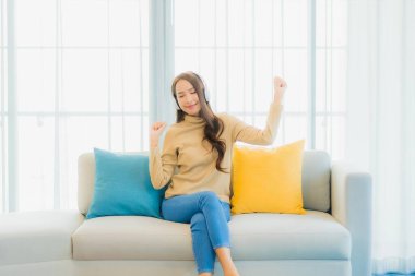 Oturma odasındaki kanepede müzik dinlemek için kulaklıklı güzel Asyalı kadın portresi.