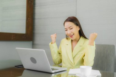 Asyalı güzel bir kadının portresi iç odasındaki çalışma masasında çalışan akıllı cep telefonlu bir bilgisayar dizüstü bilgisayar kullanıyor.