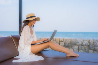 Plaj denizi çevresinde çalışmak için dizüstü bilgisayarı kullanan Asyalı genç bir kadının portresi.